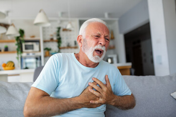 Senior man presses hand to chest has heart attack suffers from unbearable pain, Mature man with pain on heart in living room. Senior man suffering from bad pain in his chest heart attack at home