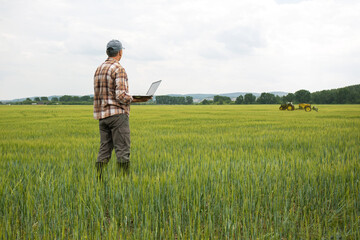  farmer standing in young wheat field with laptop and tractor spraying wheat field.