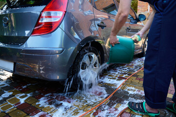Selective focus. Rear view of a man manually washing his car in the backyard, pouring water from a bucket onto the wheel © Taras Grebinets