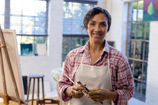 Portrait of happy biracial woman wearing apron holding brushes in home studio