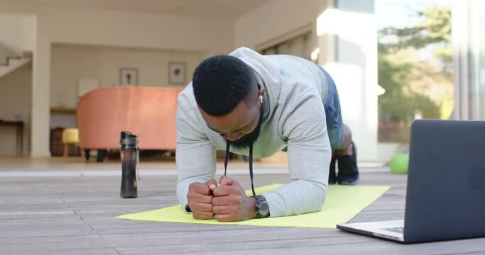 Focused african american man exercising on mat with laptop at home, slow motion