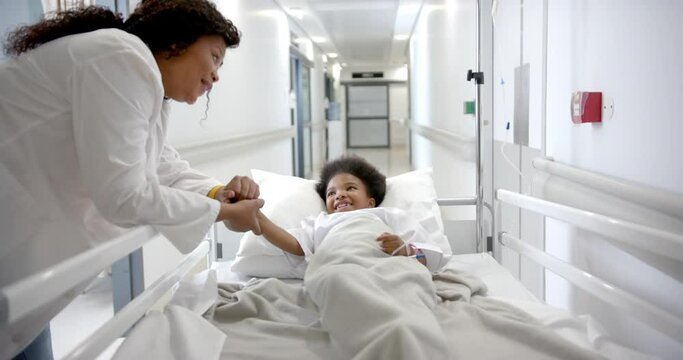 African american female doctor holding hand of girl lying in hospital bed in corridor, slow motion