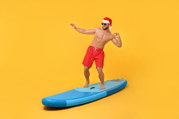 Happy man in Santa hat balancing on SUP board against orange background