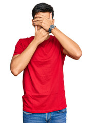 Young handsome man wearing casual red tshirt covering eyes and mouth with hands, surprised and shocked. hiding emotion