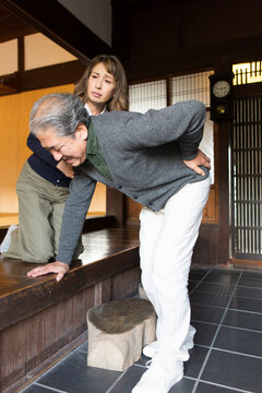 玄関で腰を押さえるシニア男性と様子を伺うシニア女性