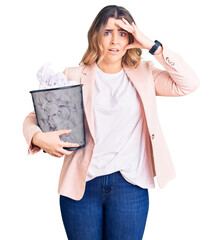 Young caucasian woman holding paper bin full of crumpled papers stressed and frustrated with hand on head, surprised and angry face