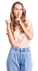 Young beautiful caucasian woman with blond hair wearing casual clothes asking to be quiet with finger on lips pointing with hand to the side. silence and secret concept.