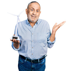 Senior man with grey hair and beard holding solar windmill for renewable electricity celebrating victory with happy smile and winner expression with raised hands