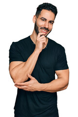 Hispanic man with beard wearing casual black t shirt looking confident at the camera with smile with crossed arms and hand raised on chin. thinking positive.