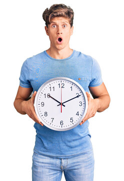 Young handsome man holding big clock scared and amazed with open mouth for surprise, disbelief face