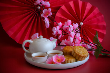 Chinese Mid-Autumn Festival concept made from mooncakes, tea decorated with plum blossom, rabbit and red paper fans on red background.