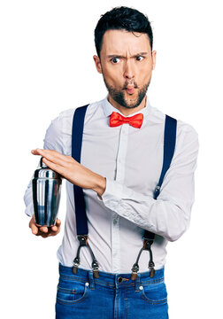 Hispanic man with beard preparing cocktail mixing drink with shaker making fish face with mouth and squinting eyes, crazy and comical.