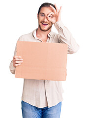 Young handsome caucasian man holding empty cardboard banner smiling happy doing ok sign with hand on eye looking through fingers