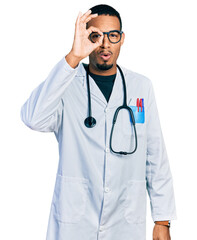 Fototapeta premium Young african american man wearing doctor uniform and stethoscope doing ok gesture shocked with surprised face, eye looking through fingers. unbelieving expression.
