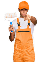 Young handsome african american man wearing cap and painter clothes holding painting roll pointing with finger to the camera and to you, confident gesture looking serious