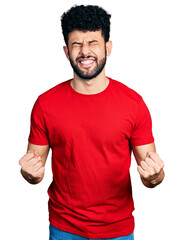 Young arab man with beard wearing casual red t shirt very happy and excited doing winner gesture with arms raised, smiling and screaming for success. celebration concept.