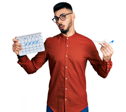 Young hispanic man with beard holding plane toy and travel calendar in shock face, looking skeptical and sarcastic, surprised with open mouth