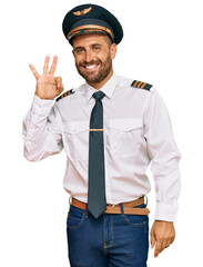 Handsome man with beard wearing airplane pilot uniform smiling positive doing ok sign with hand and...