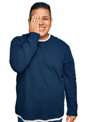 Young latin man wearing casual clothes covering one eye with hand, confident smile on face and surprise emotion.