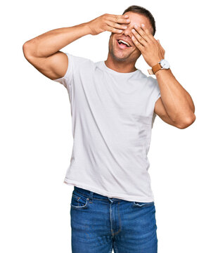 Handsome muscle man wearing casual white tshirt covering eyes with hands smiling cheerful and funny. blind concept.