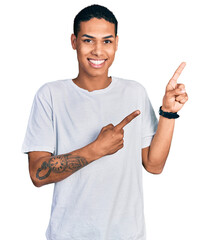 Young hispanic man wearing casual white t shirt smiling and looking at the camera pointing with two hands and fingers to the side.