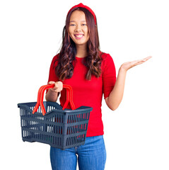 Young beautiful chinese girl holding supermarket shopping basket celebrating victory with happy smile and winner expression with raised hands