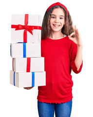Cute hispanic child girl holding gift doing ok sign with fingers, smiling friendly gesturing...