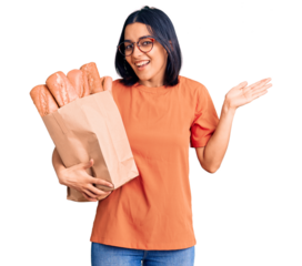 Young beautiful latin woman wearing glasses holding paper bag with bread celebrating victory with happy smile and winner expression with raised hands
