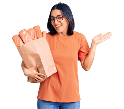 Young beautiful latin woman wearing glasses holding paper bag with bread celebrating victory with happy smile and winner expression with raised hands
