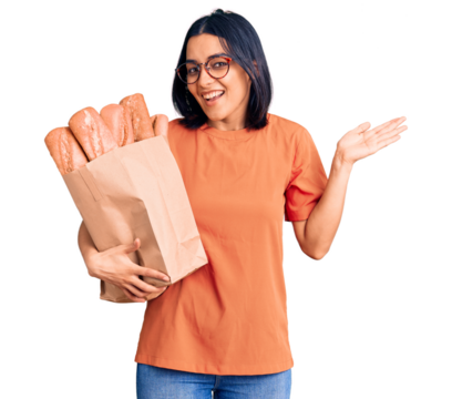 Young beautiful latin woman wearing glasses holding paper bag with bread celebrating victory with happy smile and winner expression with raised hands
