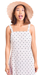 Young beautiful chinese girl wearing summer hat sticking tongue out happy with funny expression. emotion concept.