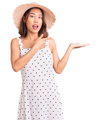 Young beautiful chinese girl wearing summer hat amazed and smiling to the camera while presenting with hand and pointing with finger.