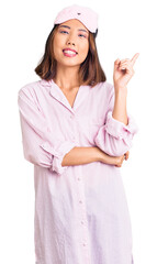 Young beautiful chinese girl wearing sleep mask and pajama with a big smile on face, pointing with hand and finger to the side looking at the camera.