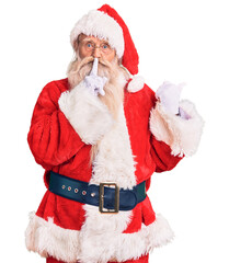 Old senior man with grey hair and long beard wearing traditional santa claus costume asking to be quiet with finger on lips pointing with hand to the side. silence and secret concept.