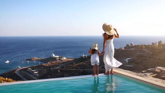 A beautiful mother woman and her daughter are standing by the pool and enjoying the summer sunset over the Aegean sea, Greece