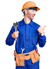 Young hispanic man wearing electrician uniform holding wrench pointing thumb up to the side smiling happy with open mouth