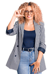 Young blonde woman with curly hair wearing business jacket and glasses smiling and confident gesturing with hand doing small size sign with fingers looking and the camera. measure concept.