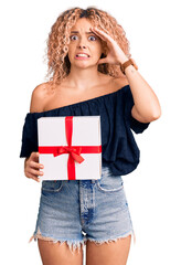 Young blonde woman with curly hair holding gift stressed and frustrated with hand on head, surprised and angry face