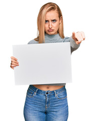 Fototapeta premium Beautiful caucasian woman holding blank empty banner pointing with finger to the camera and to you, confident gesture looking serious