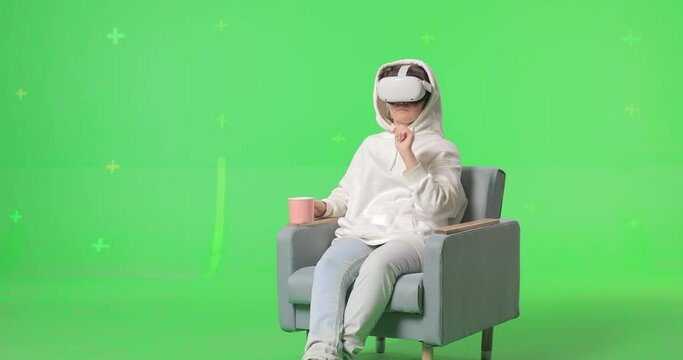 Shot on green screen background. VR or AR online at home. Cute girl in virtual reality googles in chair. Woman clicks invisible buttons pulls virtual sliders and drinking from a cup. Internet fun.