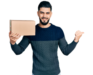 Young arab man with beard holding delivery package pointing thumb up to the side smiling happy with open mouth
