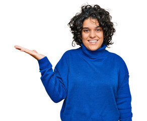 Young hispanic woman with curly hair wearing turtleneck sweater smiling cheerful presenting and pointing with palm of hand looking at the camera.