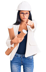 Young beautiful latin girl wearing architect hardhat holding blueprints pointing with finger to the camera and to you, confident gesture looking serious
