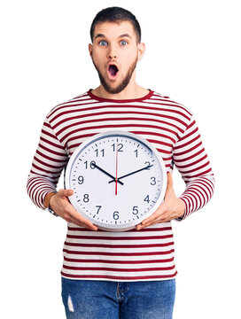 Young handsome man holding big clock scared and amazed with open mouth for surprise, disbelief face
