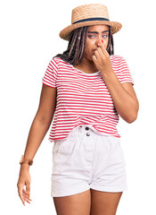 Young african american woman with braids wearing summer hat smelling something stinky and disgusting, intolerable smell, holding breath with fingers on nose. bad smell