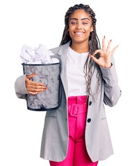 Young african american woman with braids holding paper bin full of crumpled papers doing ok sign with fingers, smiling friendly gesturing excellent symbol