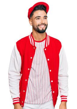 Young man with beard wearing baseball uniform looking away to side with smile on face, natural expression. laughing confident.