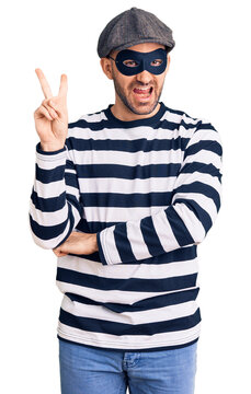 Young handsome man wearing burglar mask smiling with happy face winking at the camera doing victory sign with fingers. number two.