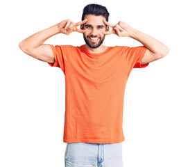 Fototapeta premium Young handsome man with beard wearing casual t-shirt doing peace symbol with fingers over face, smiling cheerful showing victory
