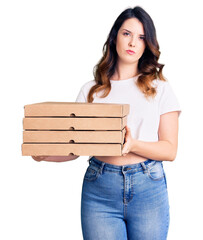 Beautiful young brunette woman holding delivery pizza box thinking attitude and sober expression looking self confident